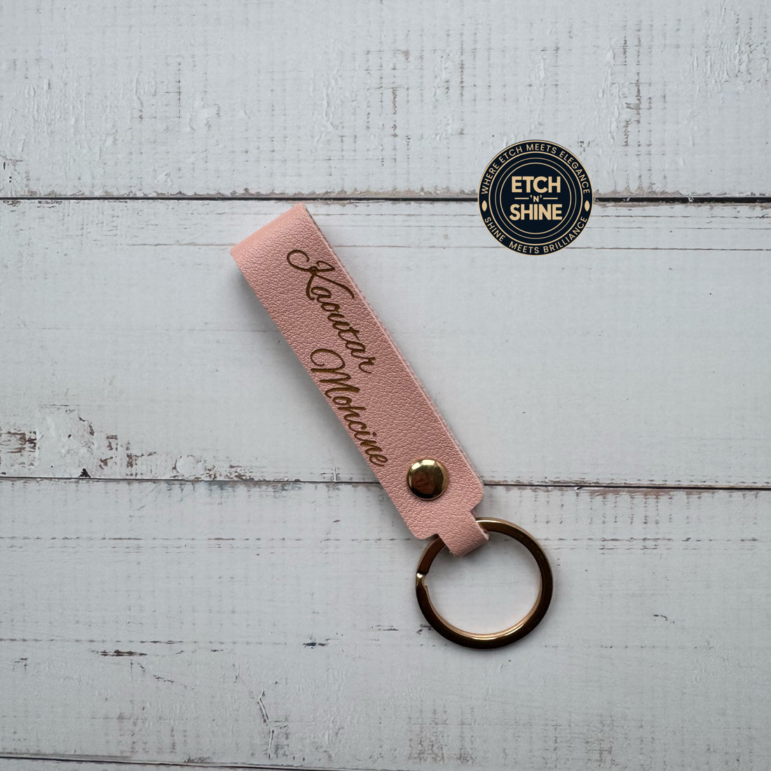 Pink keychain with engraved text on a white wooden surface, featuring the 'Etch Shine' brand.