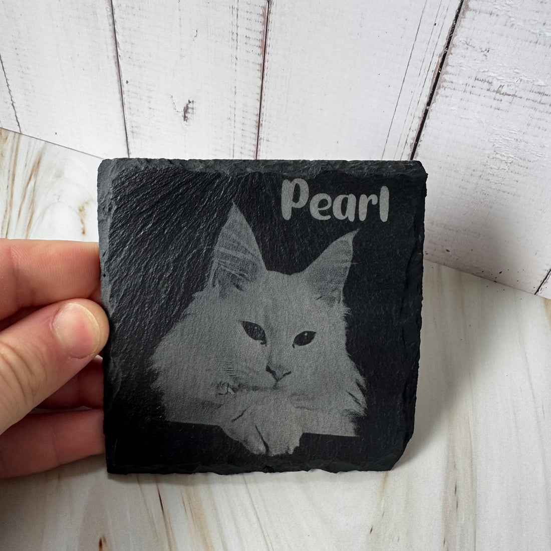 Slate coaster with engraved cat design and 'Pearl' text, held by a hand on a wooden surface.