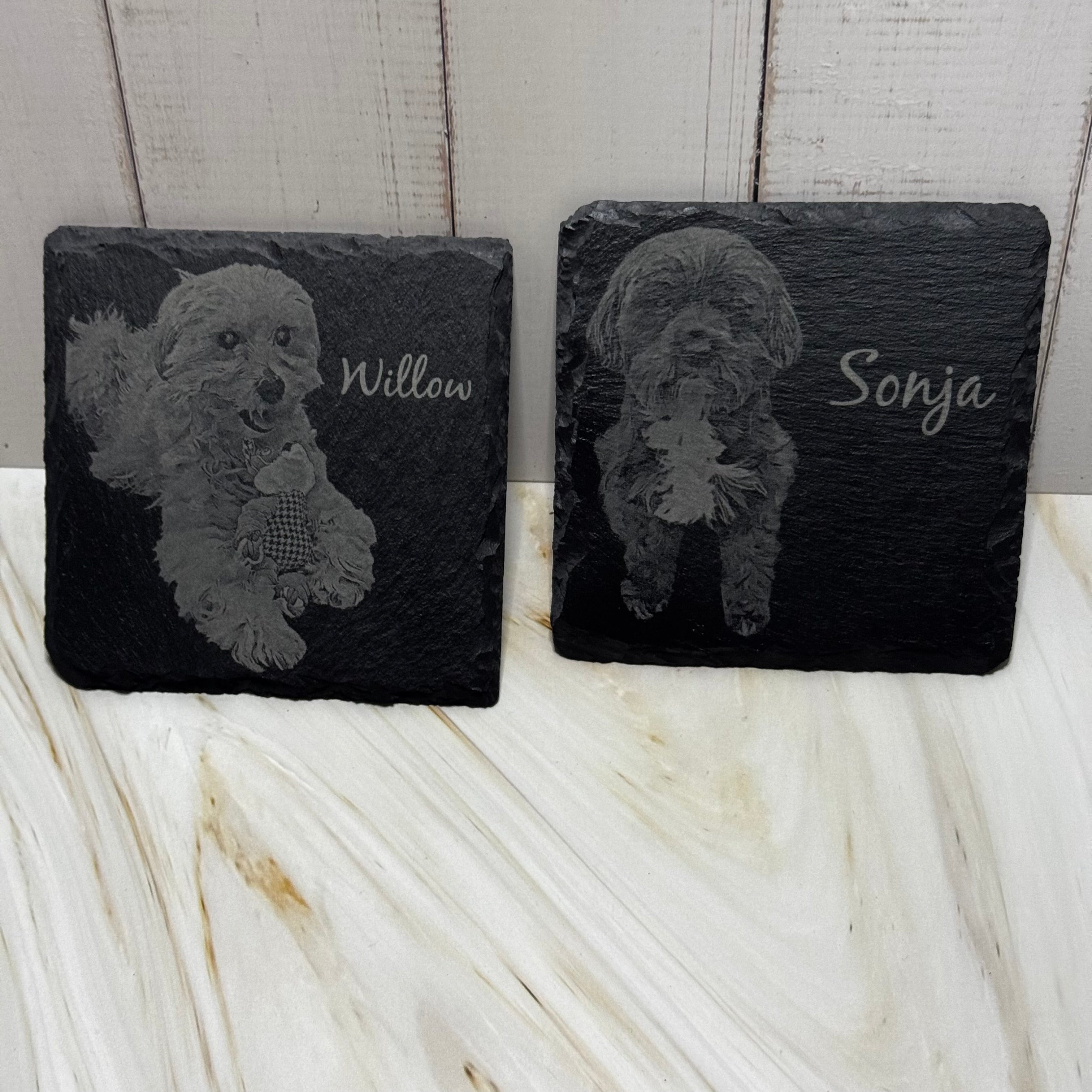 Two slate coasters with engraved dog illustrations and names on a marble surface.