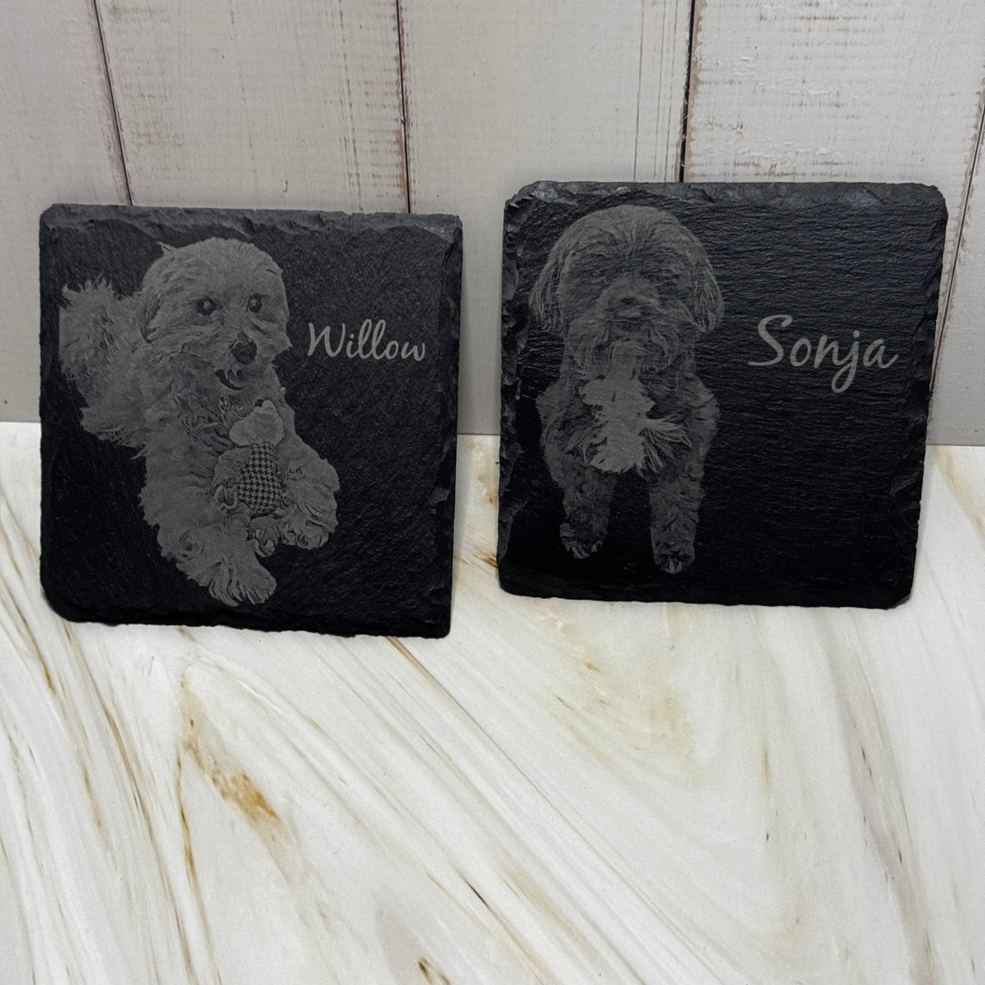 Two slate coasters with engraved dog illustrations and names on a marble surface.