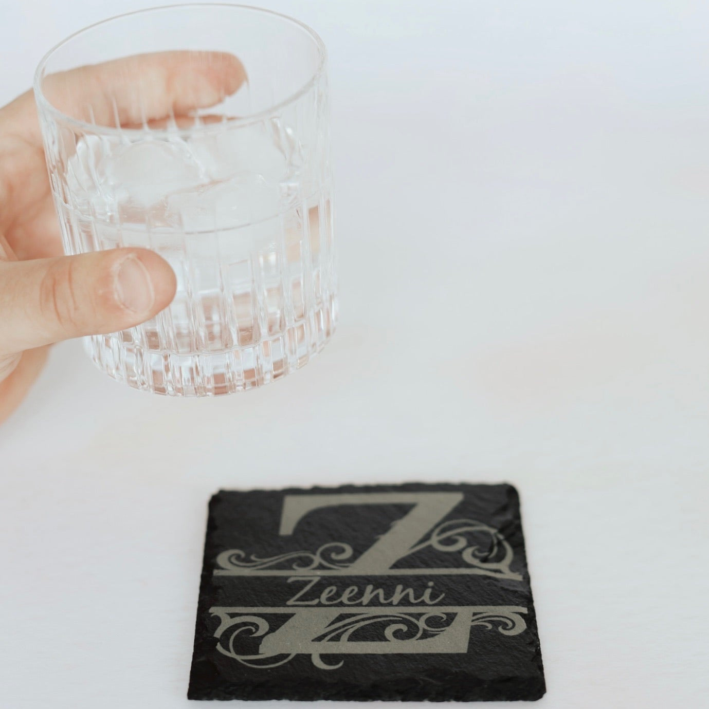 Hand holding a glass with ice on a coaster featuring the brand 'Zeeni'.