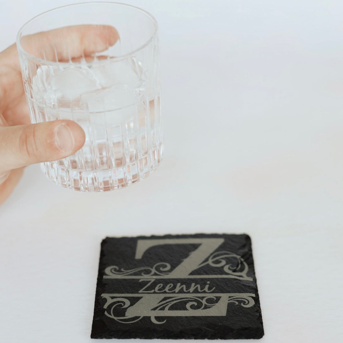 Hand holding a glass with ice on a coaster featuring the brand 'Zeeni'.