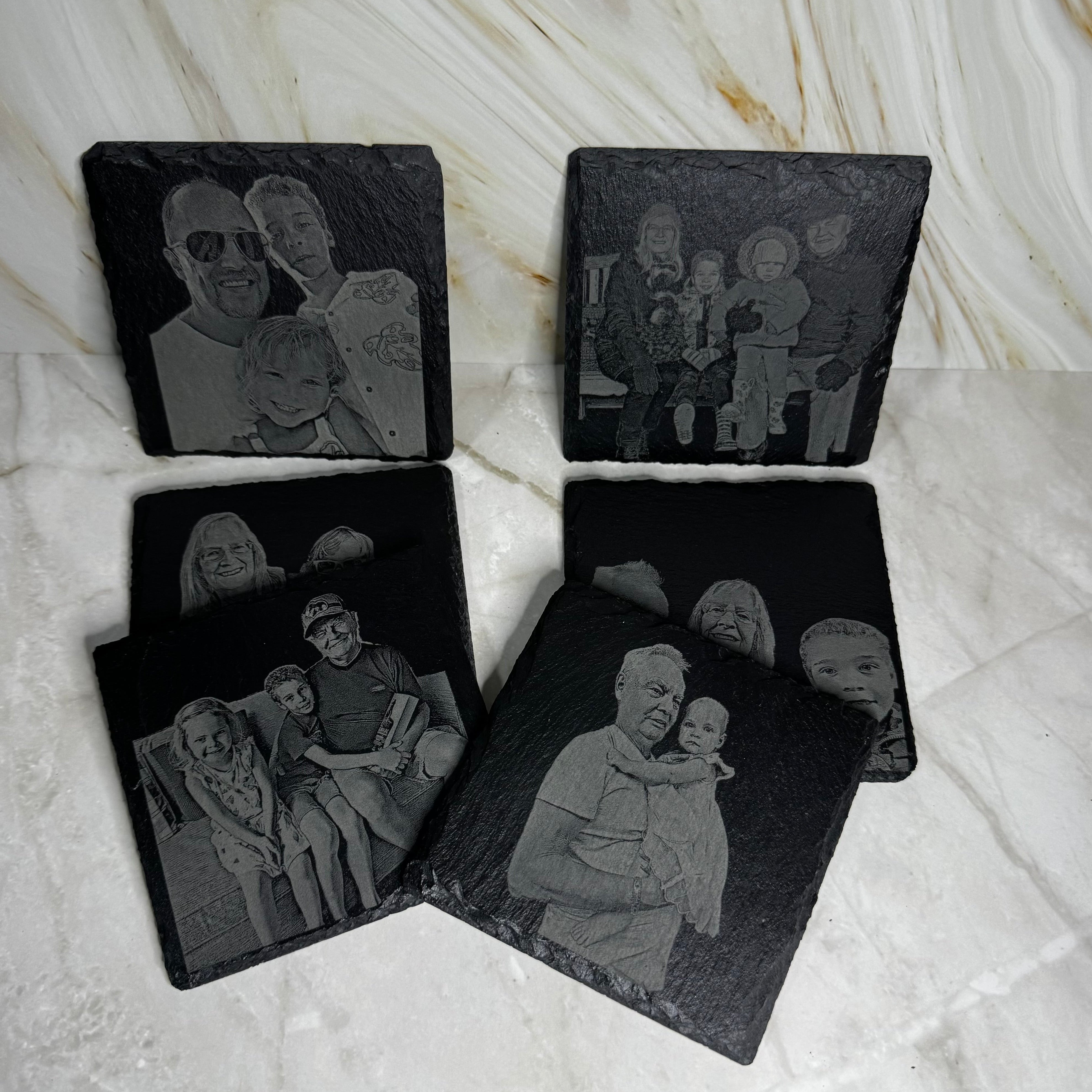 Four black slate coasters with engraved family photos on a marble surface
