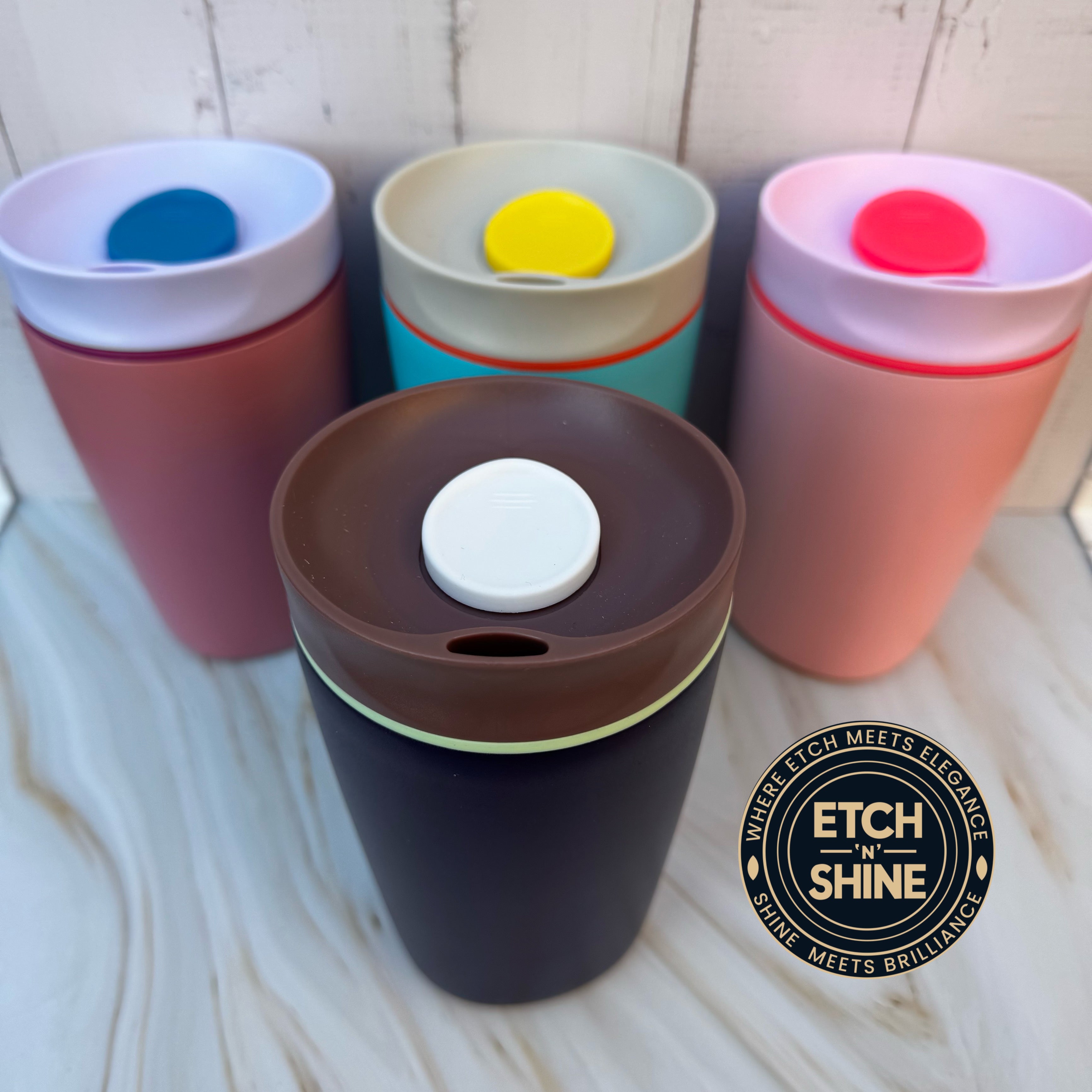 Four colorful tumblers with lids on a light wooden surface, featuring the 'Etch n Shine' brand.